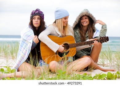 Three attractive young women sitting on the beach, wearing hats and bundled up against the cold. One woman is playing a guitar. Horizontal shot. - Powered by Shutterstock