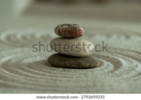 Three balanced pebbles stacked on a textured sand surface with circular ripples, creating a calm zen meditation and wellness concept.