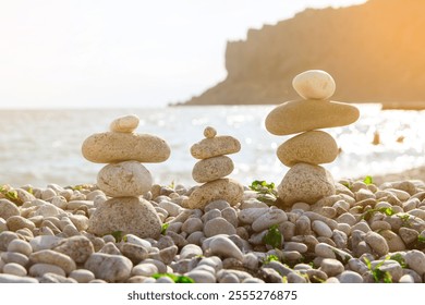 Three balanced stone stacks on a pebbly beach with a sea and rocky cliff in the background. The warm sunlight enhances the peaceful and serene atmosphere of this coastal scene - Powered by Shutterstock