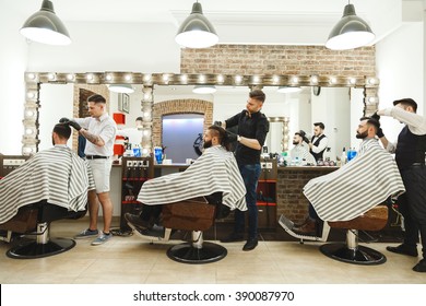 Three barbers wearing black gloves doing haircuts with scissors for men with dark hair, big mirrors next to men, three lamps over clients, portrait.
