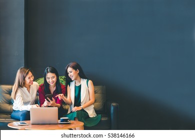 Three Beautiful Asian Girls Using Smartphone And Laptop, Chatting On Sofa Together At Cafe With Copy Space, Modern Lifestyle With Gadget Technology Or Working Woman On Casual Business Concept