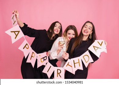 Three Beautiful Elegant Women Celebrate Birthday Party And Drinking Cocktails. Best Friends Wearing Black And White Stylish Evening Outfits With Garland With Happy Birthday Inscription. Indoors Party