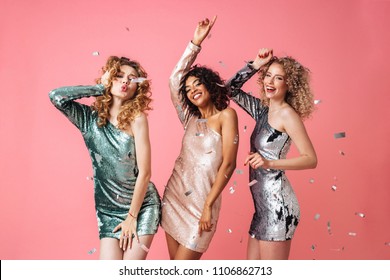 Three Beautiful Excited Women In Shiny Dresses Dancing Under Confetti Rain Isolated Over Pink Background