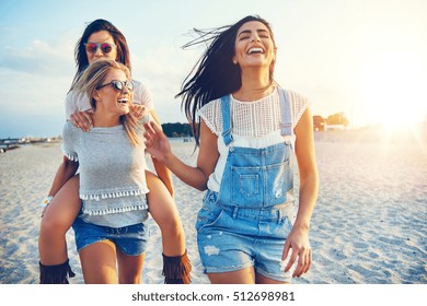 Three beautiful female friends having fun while carrying each other on beach at sunset - Powered by Shutterstock