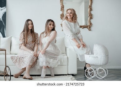 Three Beautiful Girls In Long White Dresses Are Sitting On A White Sofa At Home. Stylish White Baby Bed. Mom With Two Daughters, In The Family Circle.