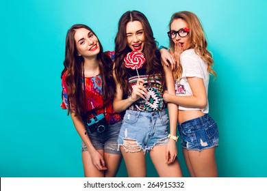 Three beautiful happy sexy women in stylish trendy  summer outfit posing against  blue  wall .Pretty girl holding   lolly-pop and her friends wearing stylish funky sunglasses and retro film camera.