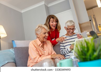 Three Beautiful Senior Women Relaxing On The Living Room Floor Looking At A Laptop Computer And Laughing At Something On The Screen. Smiling Senior Friends Looking At Laptop Together At Home On Couch