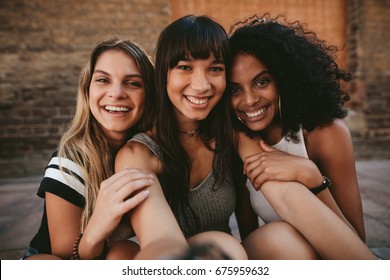 Three beautiful smiling girlfriends taking selfie with mobile phone. Multi ethnic group of women sitting outdoors by the city street and taking self portrait.