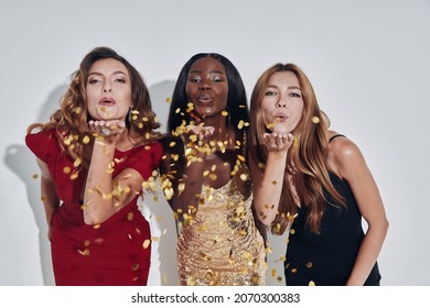 Three Beautiful Women In Evening Gowns Blowing Confetti And Smiling While Standing Against Gray Background