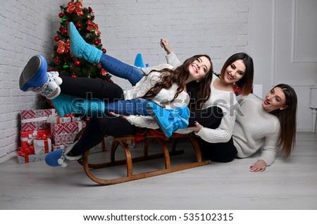 Three beautiful young girls with Christmas winter sleigh in the decorative interior of the New Year with a festive Christmas tree and Christmas decorations. Girls drop out of the sleigh.