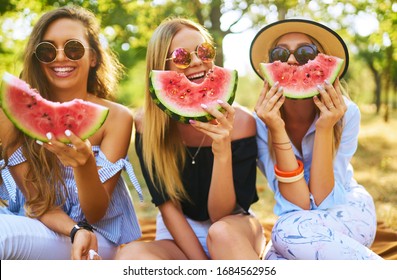 Three beautiful young girls have fun together and eating watermelon  in hot summer day. Friends holding slice of watermelon and posing in the park. Summer concept.