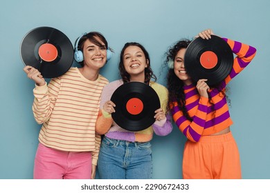 Three beautiful young girls in headphones and colorful clothes posing with vinyl records and smiling isolated over blue studio background - Powered by Shutterstock