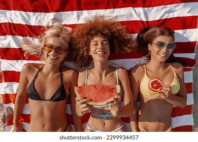 Three beautiful young women lying on the beach sunbathing in swimming suites, eating fruit, relaxing and enjoying. - Powered by Shutterstock