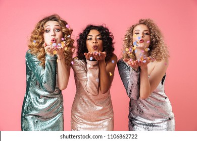 Three Beautiful Young Women In Shiny Dresses Blowing Confetti From Their Palms Isolated Over Pink Background