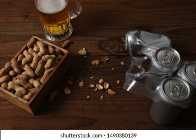 Three Beer Cans In A Plastic 6-pack Holder A Crushed Can And A Mug Of Beer With A Wood Box Of Peanuts. Used Shells Litter The Rustic Dark Wood Table.