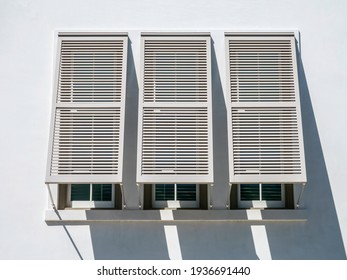 Three Bermuda shutters propped open by three windows of building with whitewashed wall on a sunny morning in the Florida Panhandle