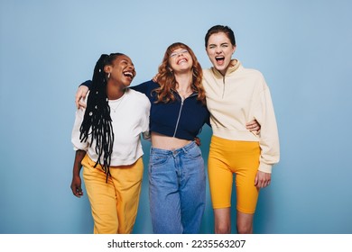 Three best friends laughing and having a good time while embracing each other in a studio. Group of happy female friends enjoying themselves while standing against a blue background.