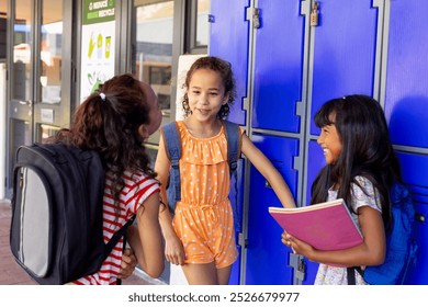 Three biracial girls chat by school lockers, one holding a pink folder. They wear casual clothes, capturing a moment of friendship and school life. - Powered by Shutterstock