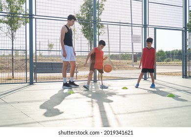 Three Brothers Playing Basketball. Older Brother Teaching How To Play Basketball To His Younger Siblings, One Of The Kids Has A Leg Prosthesis.