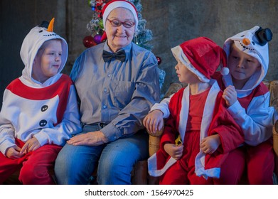 Three brothers in xmas clothes play with tablet and smartphone. Granny sits next to her grandsons and looks at their entertainment. Beautiful elderly woman with gray hair and wrinkles. Kids love mom