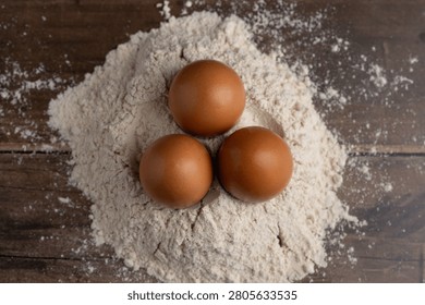 Three brown eggs rest together in a soft nest of scattered flour on a rustic wooden surface, evoking themes of baking and food preparation.