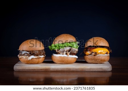 Three burger sliders placed on a wooden board. Very tasty burgers with melted cheese, cheddar, bacon, lettuce, tomato, onion and sauce. Craft beef burgers, hand made.