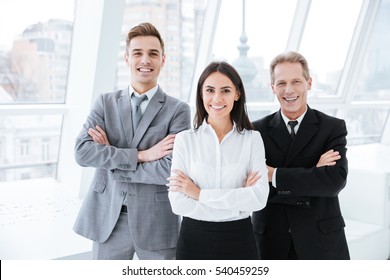 Three Business People With Arms Crossed Standing Near The Window In Conference Room