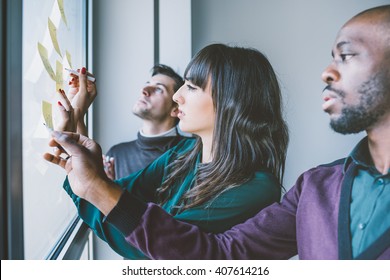 Three business people having a meeting in office. They are standing in front of glass wall with post it notes, pointing and discussing - business, teamwork, brainstorming concept