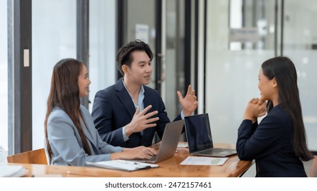 Three business professionals engaged in a meeting around a table, discussing projects with laptops in an office setting. - Powered by Shutterstock