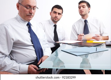 Three businessman, one mature texting using his mobile phone and two young ones sitting at table during meeting