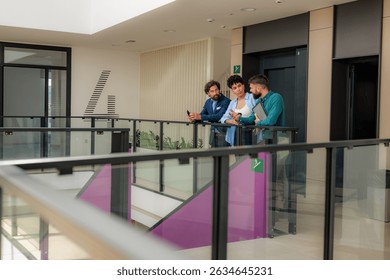 Three businesspeople are having a conversation about work while leaning on a railing in a modern office building, with one holding a tablet and another holding a phone