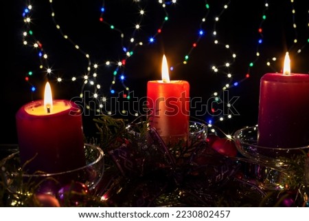 Three candles glow on Advent Wreath. Lighting the third candle on the third Sunday in December. Festive red, blue and golden holiday bokeh background. Celebrating the beginning of Christmas holidays.