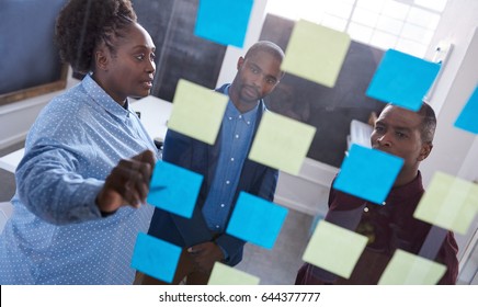 Three casually dressed African work colleagues brainstorming together with sticky notes on a glass wall while standing in a modern office