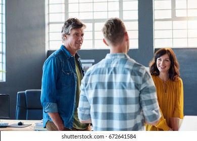Three casually dressed young office coworkers smiling and talking together while standing in a large modern office