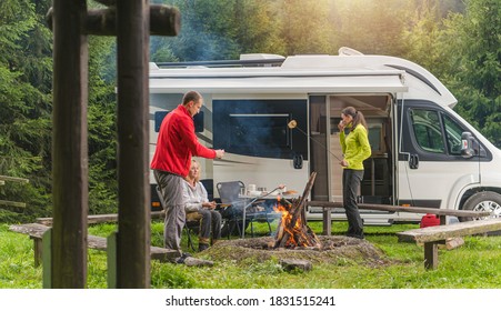 Three Caucasian Friends In Their Late 30s, One Men And Two Woman Having Fun On Wilderness Camping With Modern RV Camper Van. Grilling Bread And Polish Sausage On Campfire.