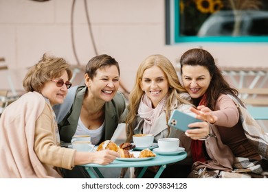 Three Caucasian Pretty Senior Women Friends Sitting With Coffee At Table In Cafe Terrace And Smiling To Smartphone Camera While Taking Selfie Photos. Old Ladies Posing To Phone. Pensioners Resting.
