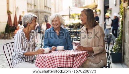 Three Caucasian senior women friendly chatting and drinking coffee at table in cafe terrace in summer. Female friends talking and laughing outdoor while gossiping and sipping tea.