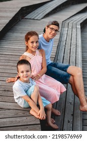 Three Caucasoid Teenagers From The Same Family Of Different Ages Sister With Two Brothers On Wooden Benches Together Side By Side Smiling At Sunset In Summer With Bare Feet