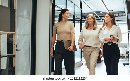 Three cheerful businesswomen walking together in an office. Diverse group of businesswomen smiling while having a discussion. Successful female colleagues collaborating on a new project. - Shutterstock ID 2112192284