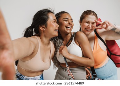 Three cheerful diverse women in sportswear and with yoga mat taking selfie isolated over white background - Powered by Shutterstock