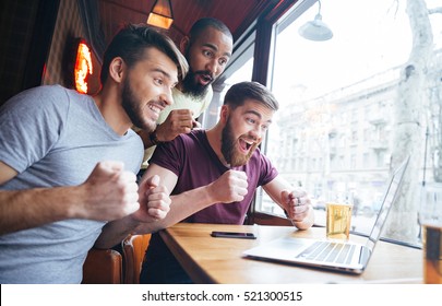 Three cheerful excited young men watching match on laptop and supporting their team sitting in bar