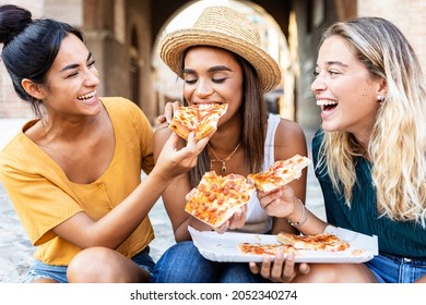 Three cheerful multiracial women eating pizza in the street - Happy millennial friends enjoying the weekend together while sightseeing an italian city - Young people lifestyle concept