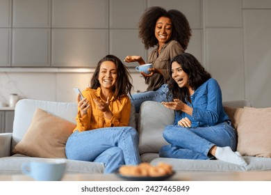 Three cheerful multiracial women friends share laughter in living room, one holding mobile phone, enjoying their coffee and relaxing weekend together, sitting on sofa at home interior - Powered by Shutterstock