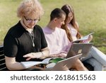 Three cheerful students study and work together outdoors on a sunny day, using laptops and notebooks while sitting on a park bench