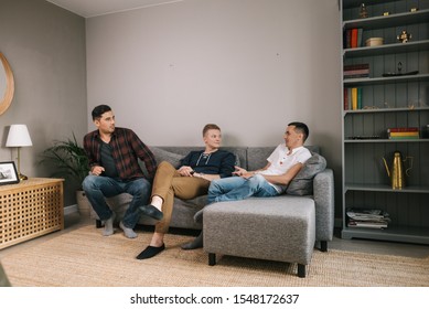 Three Cheerful Young Guys Are Sitting On Soft Couch At Home And Have Fun Conversation. Group Of Friends Are Sitting On A Soft Couch And Communicates Carefreely In Living Room With Modern Interior.