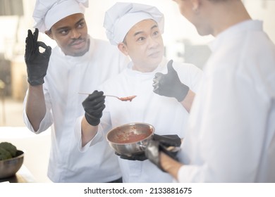 Three Chef Cooks With Different Ethnicities Tasting Sauce With A Spoon While Cooking In The Kitchen. Teamwork And Cooking Delicious Food At Restaurant