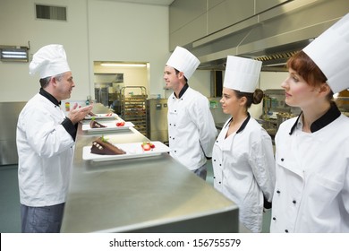 Three chefs presenting their dessert plates to the head chef in busy kitchen - Powered by Shutterstock