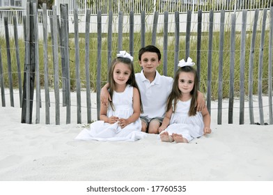 Three Children, 2 Girls And 1  Boy Sitting On The Beach By A Fence Dressed In White.