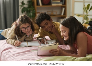 Three children of diverse backgrounds studying and smiling together while lying on bed with books and notebooks surrounding them, creating an engaging and lively atmosphere - Powered by Shutterstock