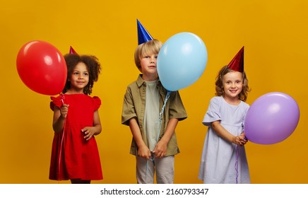 Three children in festive hats on their heads with colorful balloons in their hands in anticipation of the holiday and emotions.